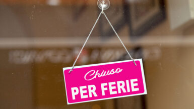 Red sign hanging at the glass door of a shop saying in Italian "Chiuso per ferie", meaning in English "Closed for holiday".